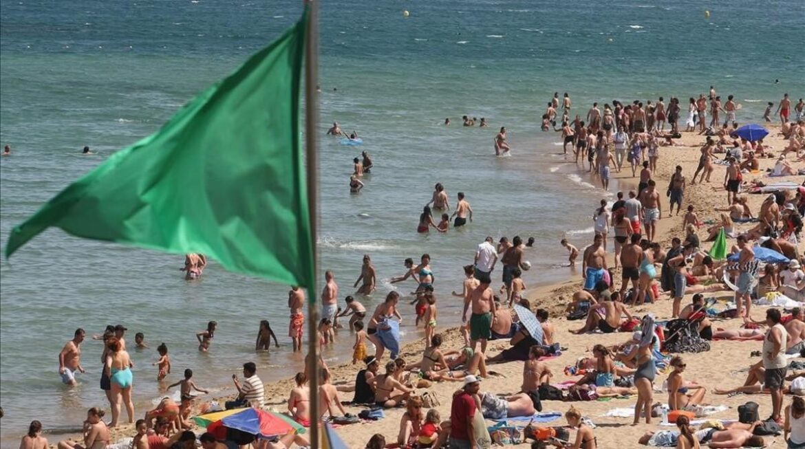 Playa concurrida con bañistas y bañistas, bandera verde y mar tranquilo al fondo.